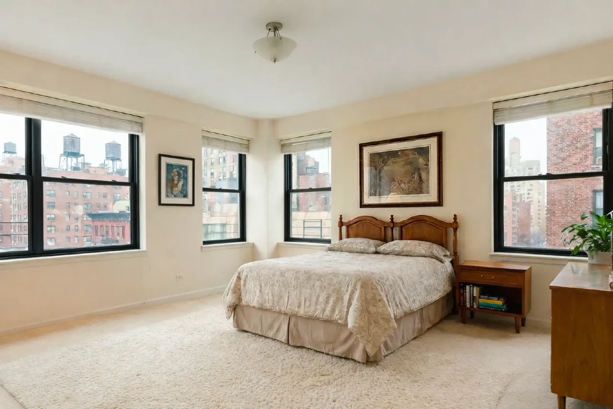**Bright bedroom interior featuring beige carpeting, wooden furniture, and multiple windows with a city view.**