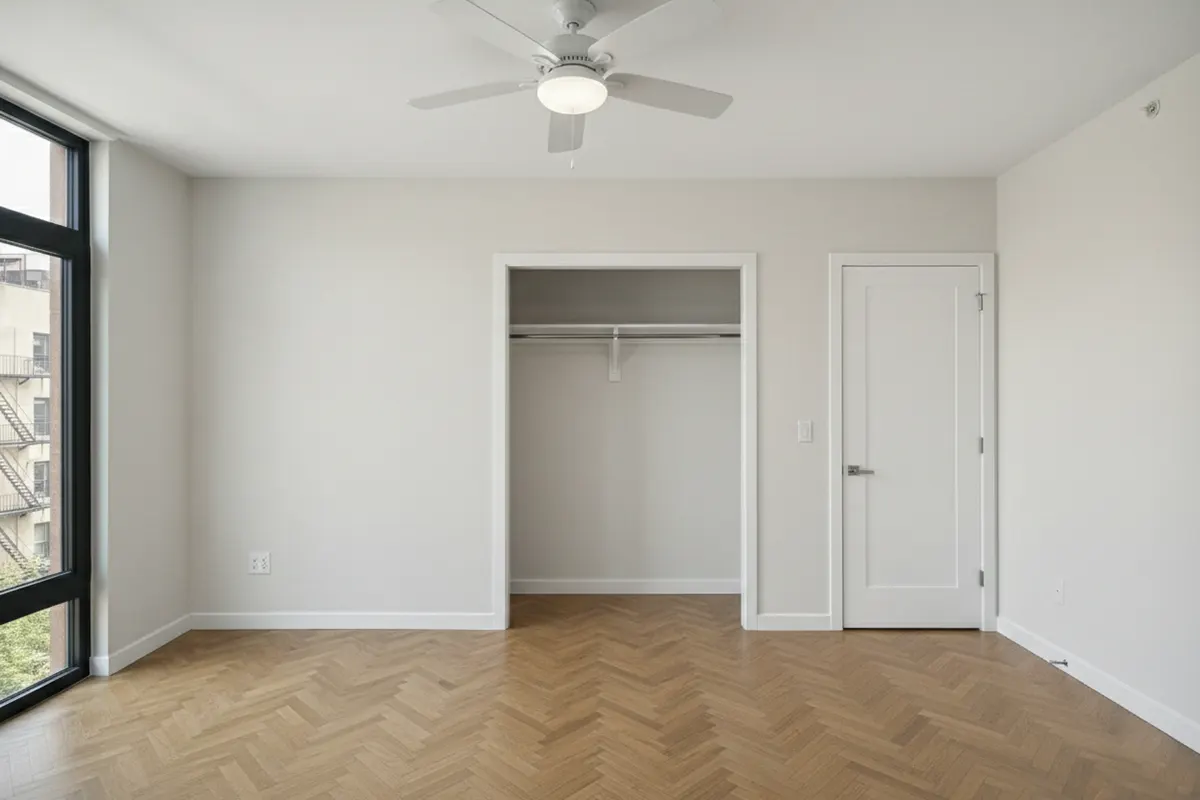 Empty New York apartment bedroom featuring herringbone wood floors, an open closet, a ceiling fan, and urban city views.