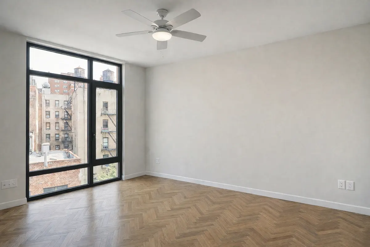 Modern empty New York bedroom featuring herringbone wood floors, a ceiling fan, and large black-framed windows with urban city views.