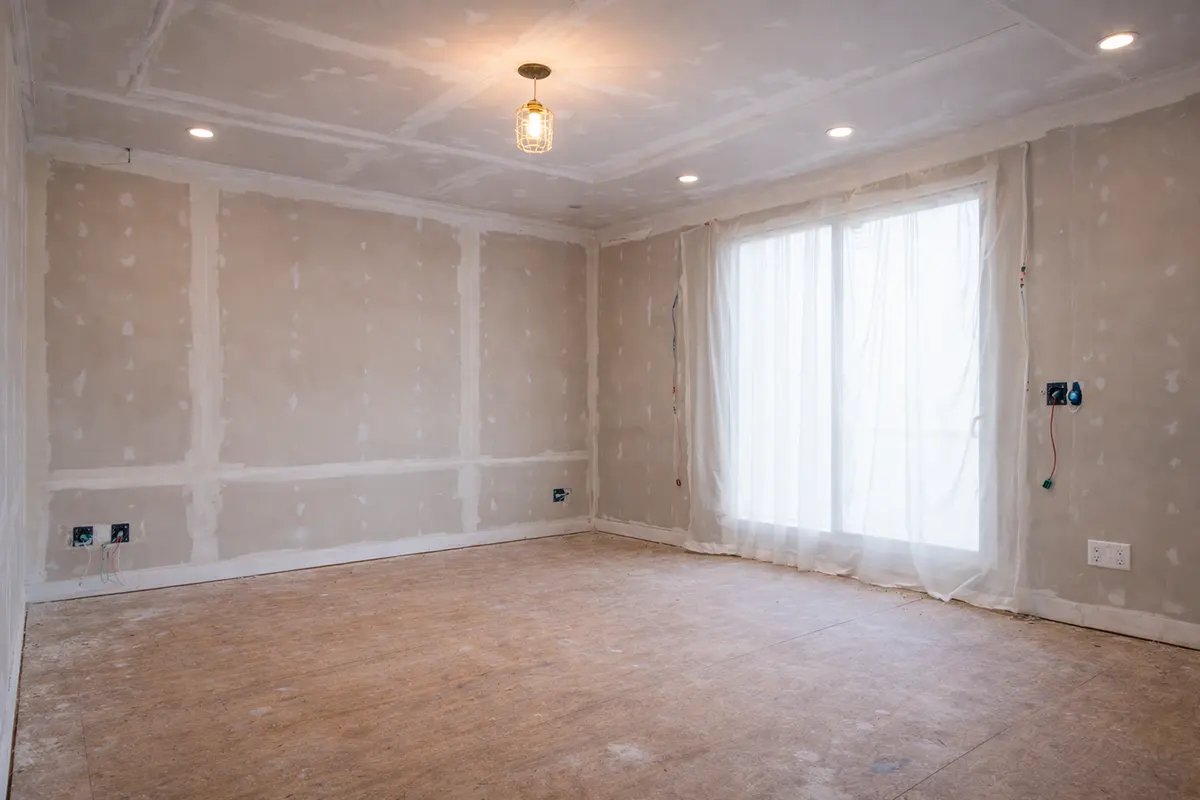 Room interior under renovation featuring unfinished drywall with taped seams, recessed ceiling lights, and a large window covered with protective plastic.