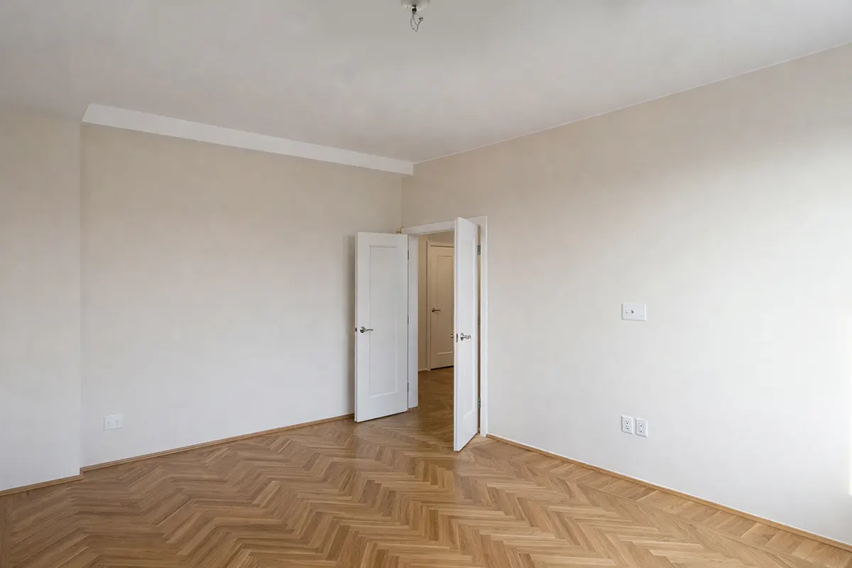 Empty Manhattan apartment room featuring herringbone wood floors, solid white double doors, and beige walls before furnishing.