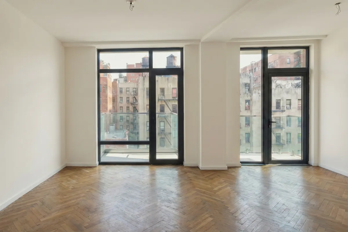 Empty modern Manhattan living room featuring herringbone wood floors, balcony access, and floor-to-ceiling windows with urban city views.