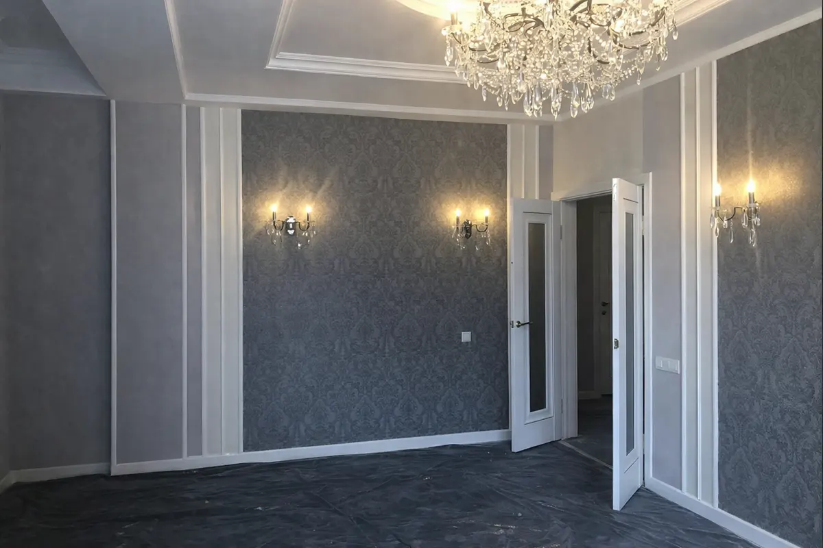 Renovated luxury living room interior featuring grey damask wallpaper panels, crystal chandeliers, and protective floor coverings.