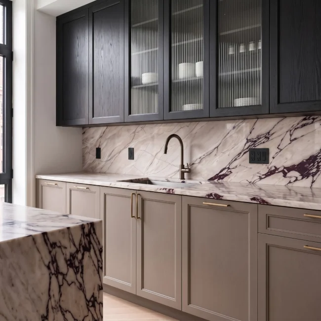 Close-up of a luxury kitchen remodel featuring ribbed glass and black upper cabinets, beige lower shaker cabinetry with brass handles, and a dramatic Calacatta Viola marble backsplash and countertop