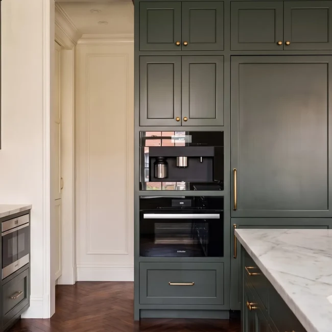 High-end kitchen remodel featuring integrated luxury appliances, a built-in coffee station and oven unit, forest green custom shaker cabinets with brass hardware, and a marble countertop