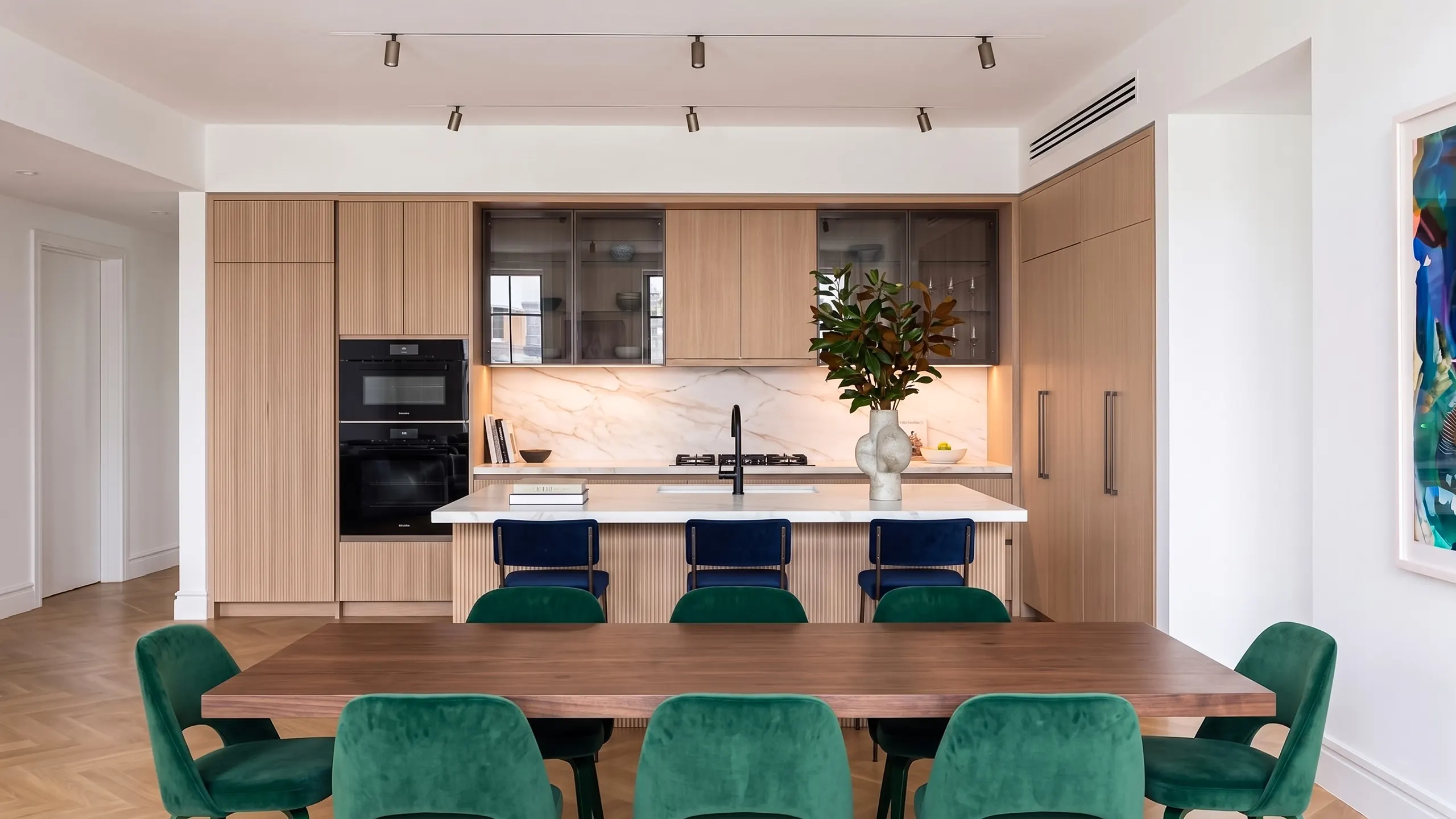 Finished luxury kitchen renovation in NYC featuring seamless light wood fluted cabinetry, a white marble kitchen island with navy blue bar stools, and a walnut dining table surrounded by green velvet chairs