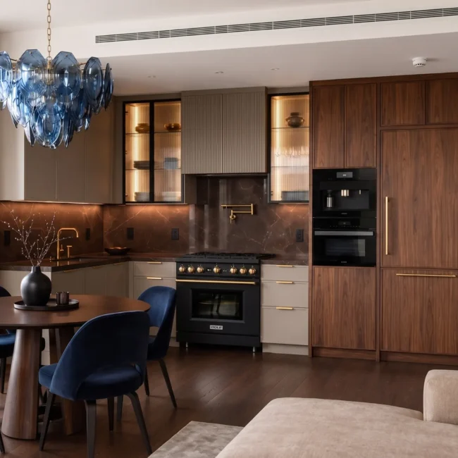 Luxury open-concept kitchen remodel in a Manhattan apartment featuring custom walnut cabinetry, dark marble backsplash, integrated appliances, and a dining area with blue velvet chairs under a statement chandelier
