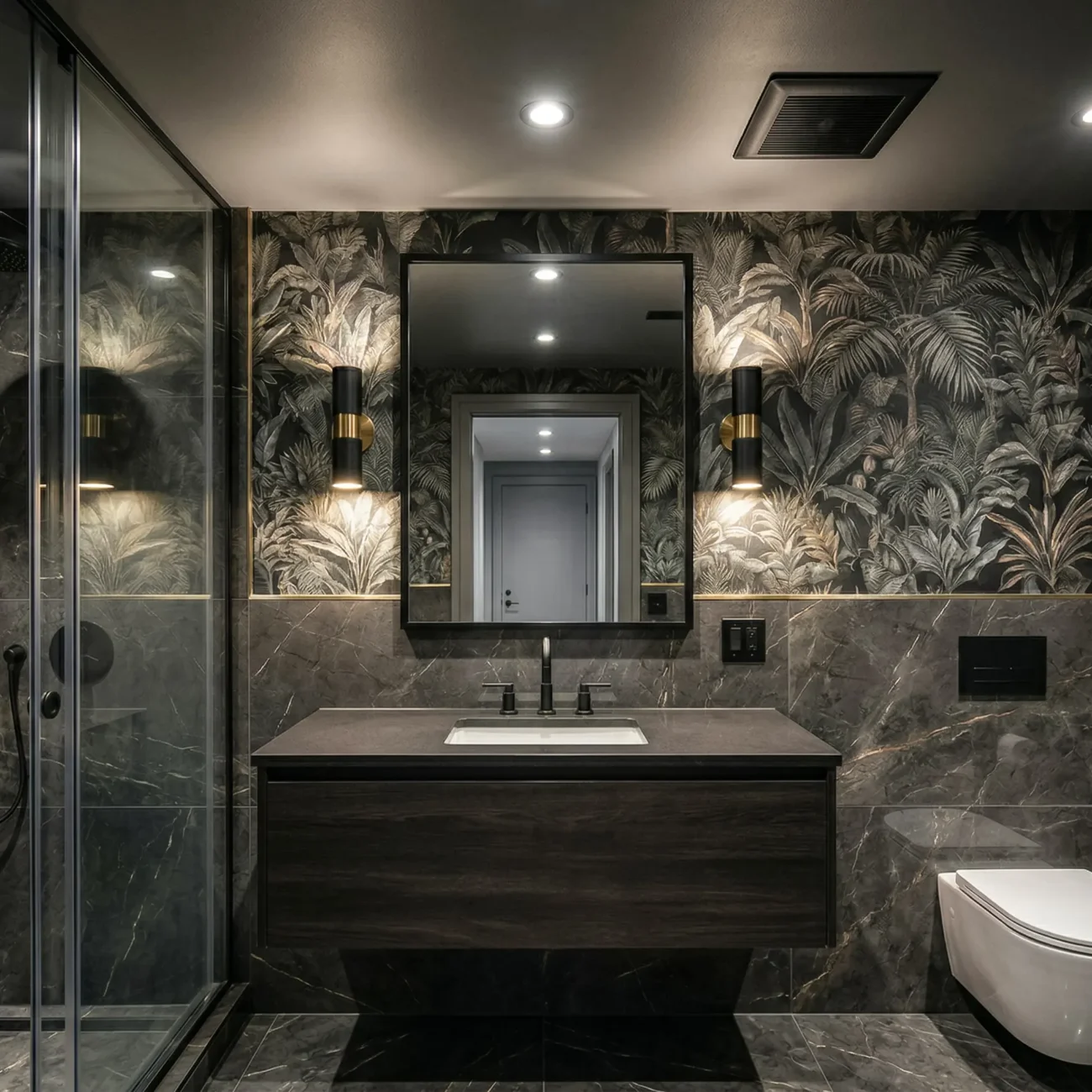 Completed small apartment bathroom remodel featuring dark marble wall tiles, a floating dark wood vanity with a black faucet, a large rectangular mirror flanked by modern black and brass sconces, a wall-mounted toilet, and a glass-enclosed walk-in shower set against a bold tropical botanical wallpaper accent