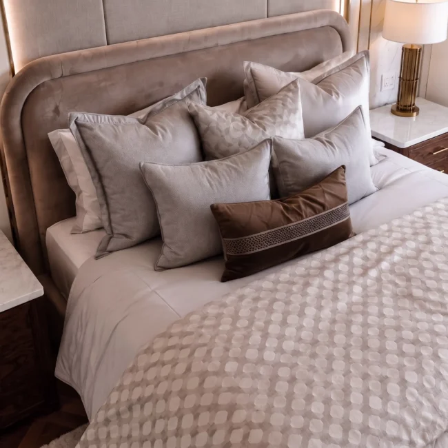 Detailed view of a luxury master bedroom remodel showing a velvet tufted headboard, premium neutral pillows and bedding, dark walnut nightstands with white marble tops, and matching columns of brass bedside lamps set against an elegant paneled wall.