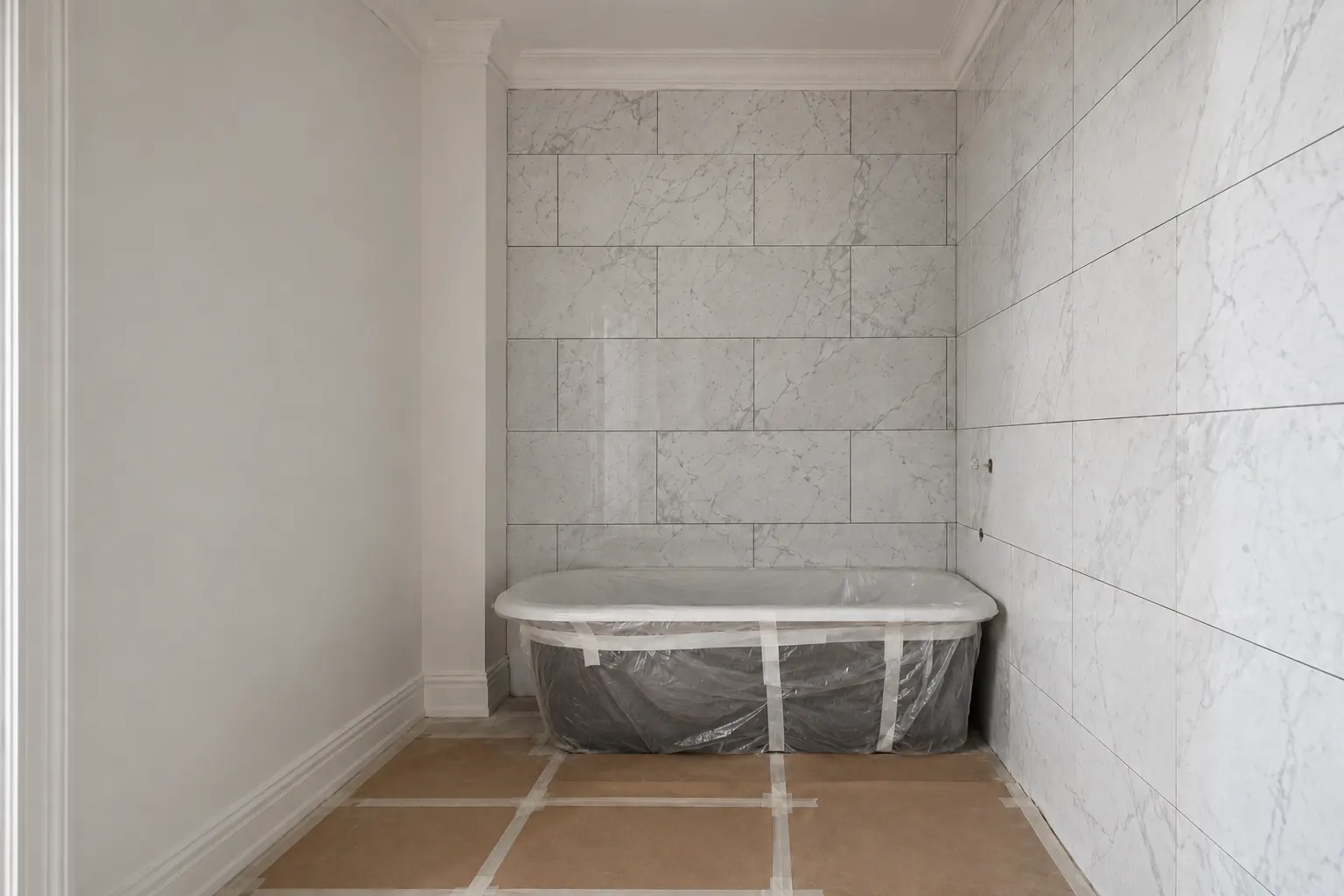 Apartment bathroom mid-renovation with white marble set on two walls and the joints still open, a bathtub wrapped in taped plastic standing against the far wall, and paper taped over the floor