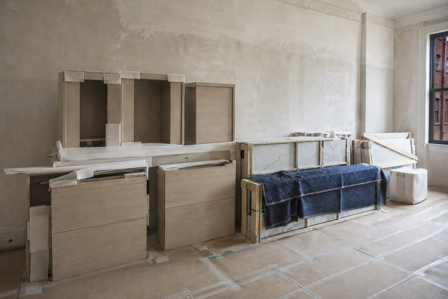  Pale oak cabinet boxes and crated marble slabs under blue moving blankets standing in a stripped pre-war apartment room with the floor covered and taped, and a window looking onto a neighboring brick building