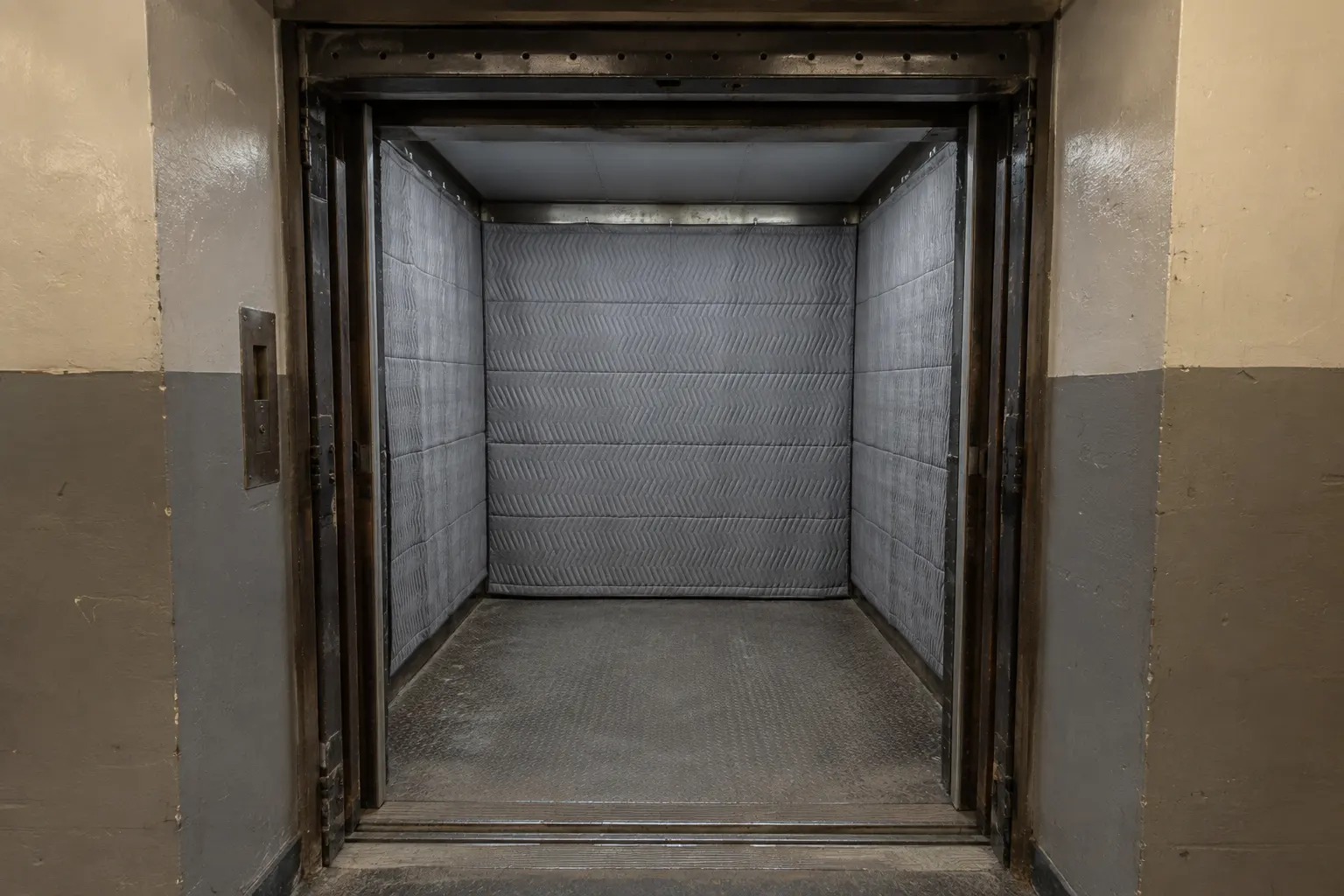 Empty freight elevator car seen from the corridor, its walls hung with quilted protective pads, with a worn steel floor and painted corridor walls on either side