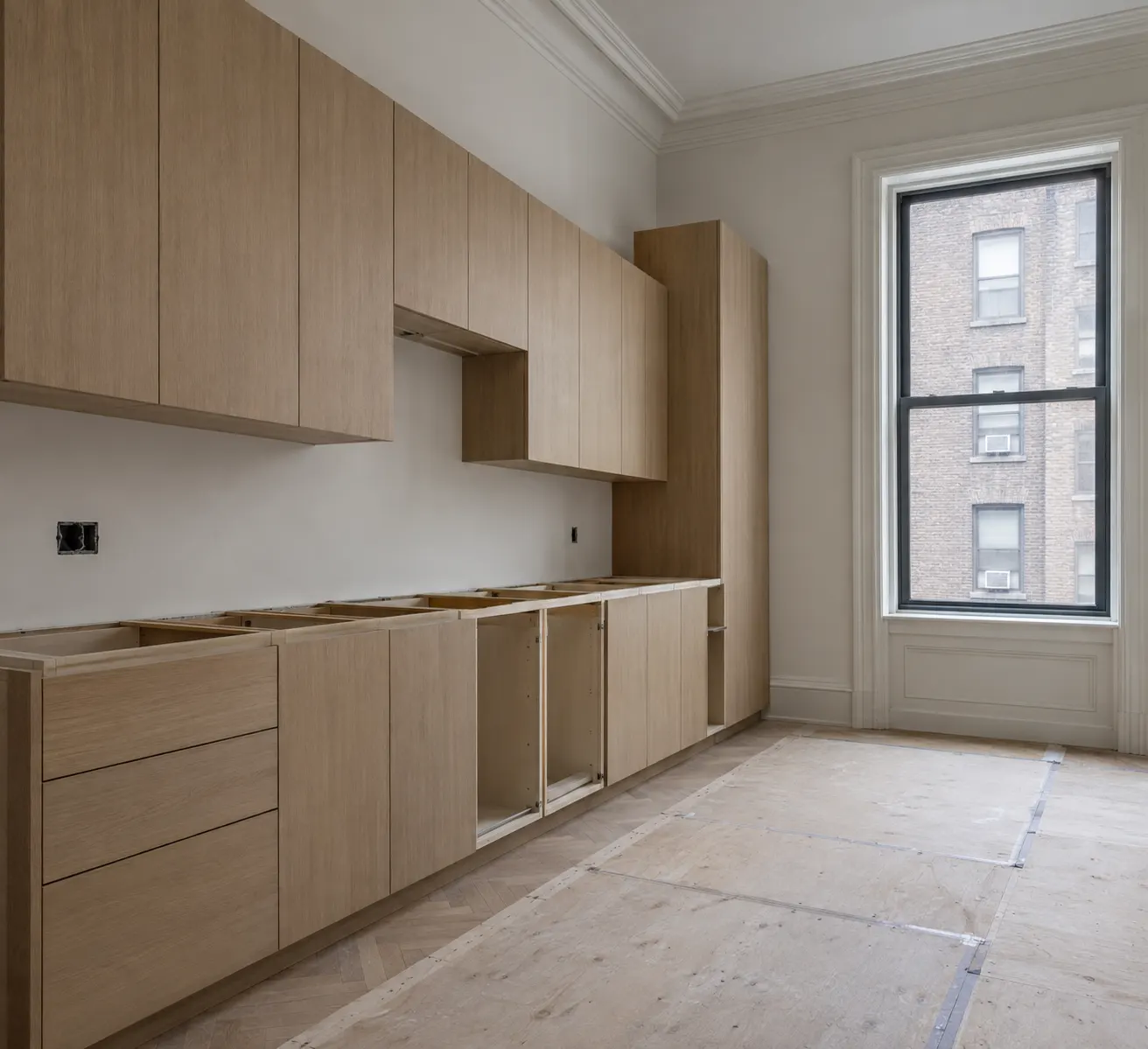 White oak kitchen cabinets fixed to the wall of a pre-war Manhattan apartment during installation, some base units still open without fronts, with rough electrical boxes on the wall and plywood protecting the herringbone floor