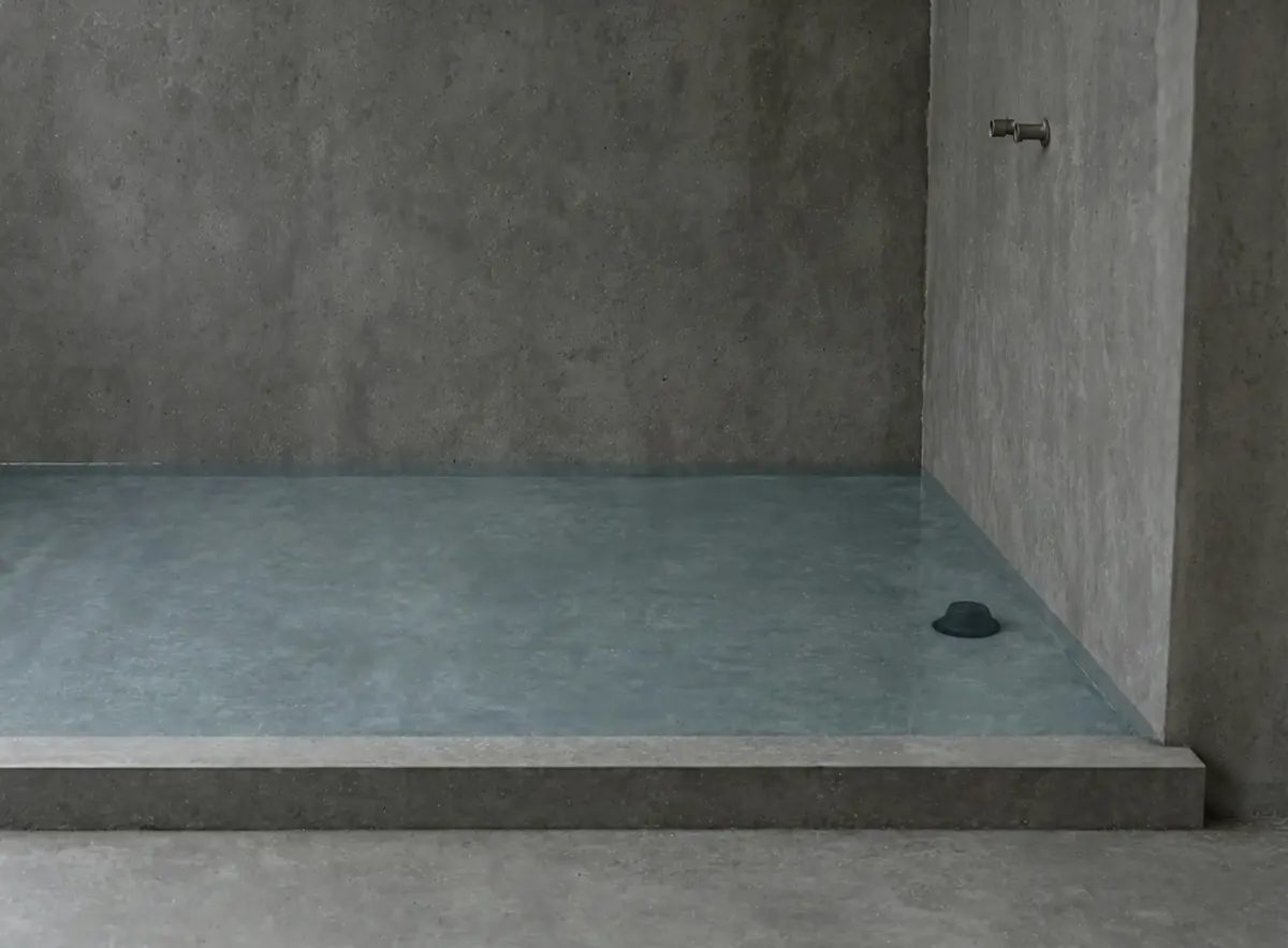Standing water held inside a shower area finished in grey waterproofing, with a dark rubber test plug in the pan and the floor beyond the low curb completely dry

