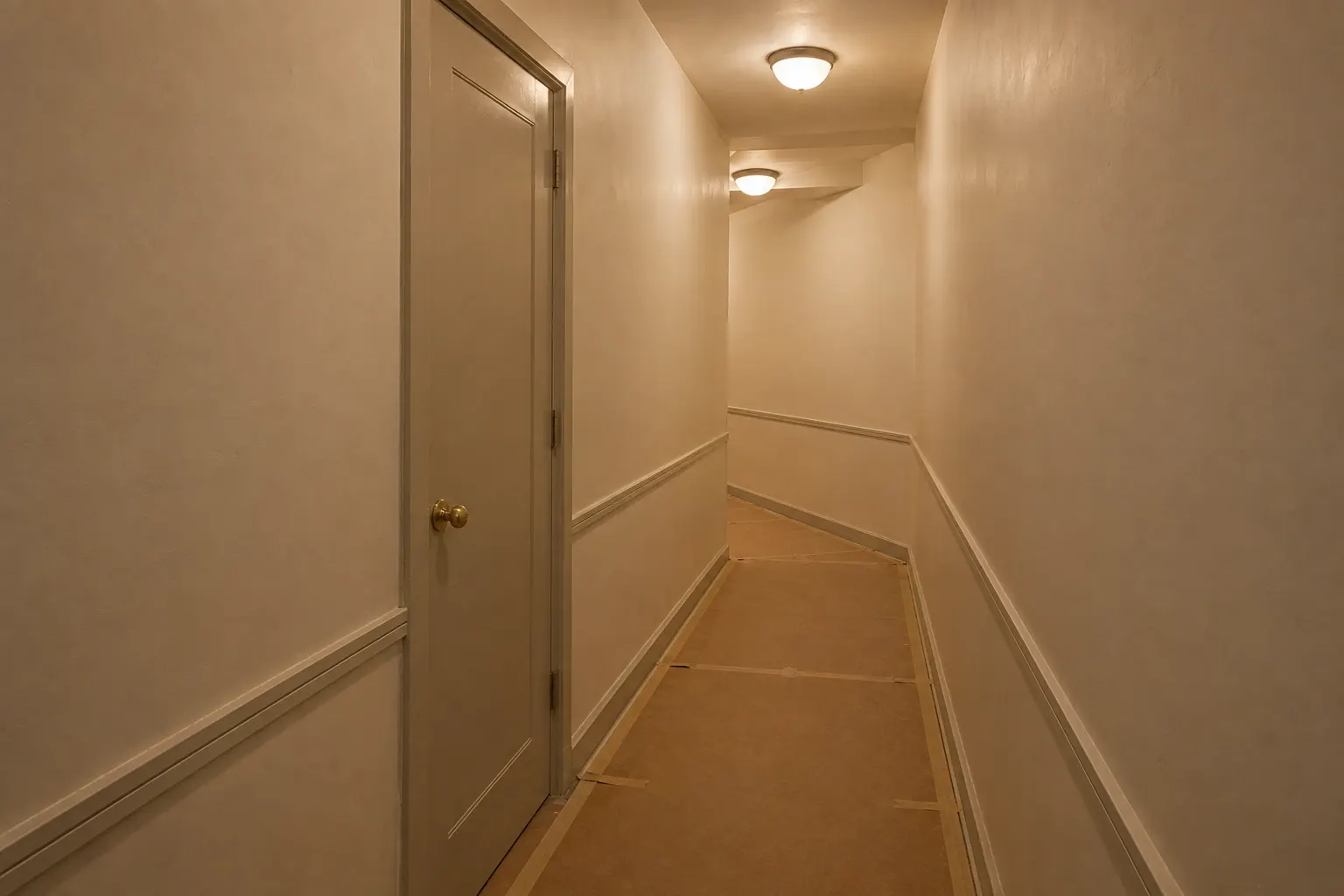 Narrow corridor in a pre-war building with brown hardboard taped over the floor, a painted apartment door with a brass knob on the left, and the corridor turning left at the far end 