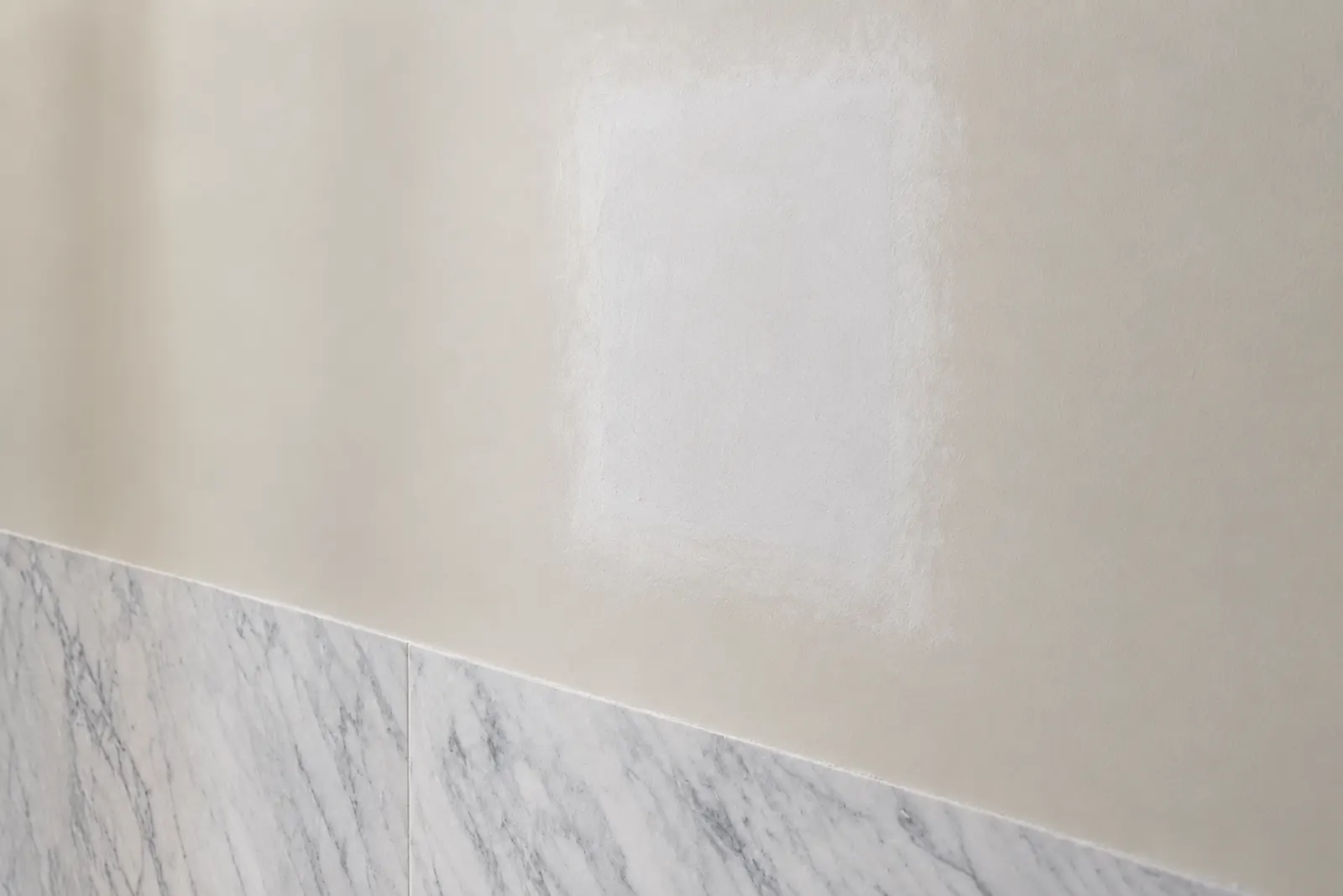 Fresh unpainted plaster patch on a pre-war bathroom wall above a Carrara marble wainscot, where the wall was opened to reach hidden wiring