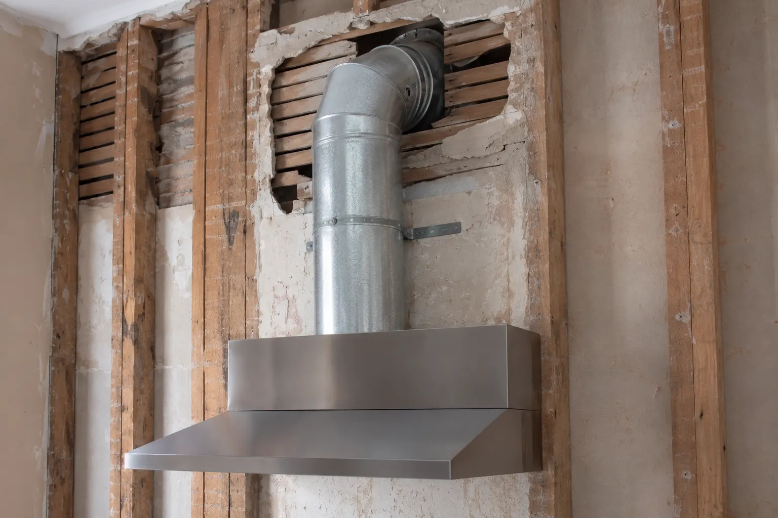 Round galvanized duct rising from a range hood housing and turning through an elbow into a wall opened down to the old plaster and lath