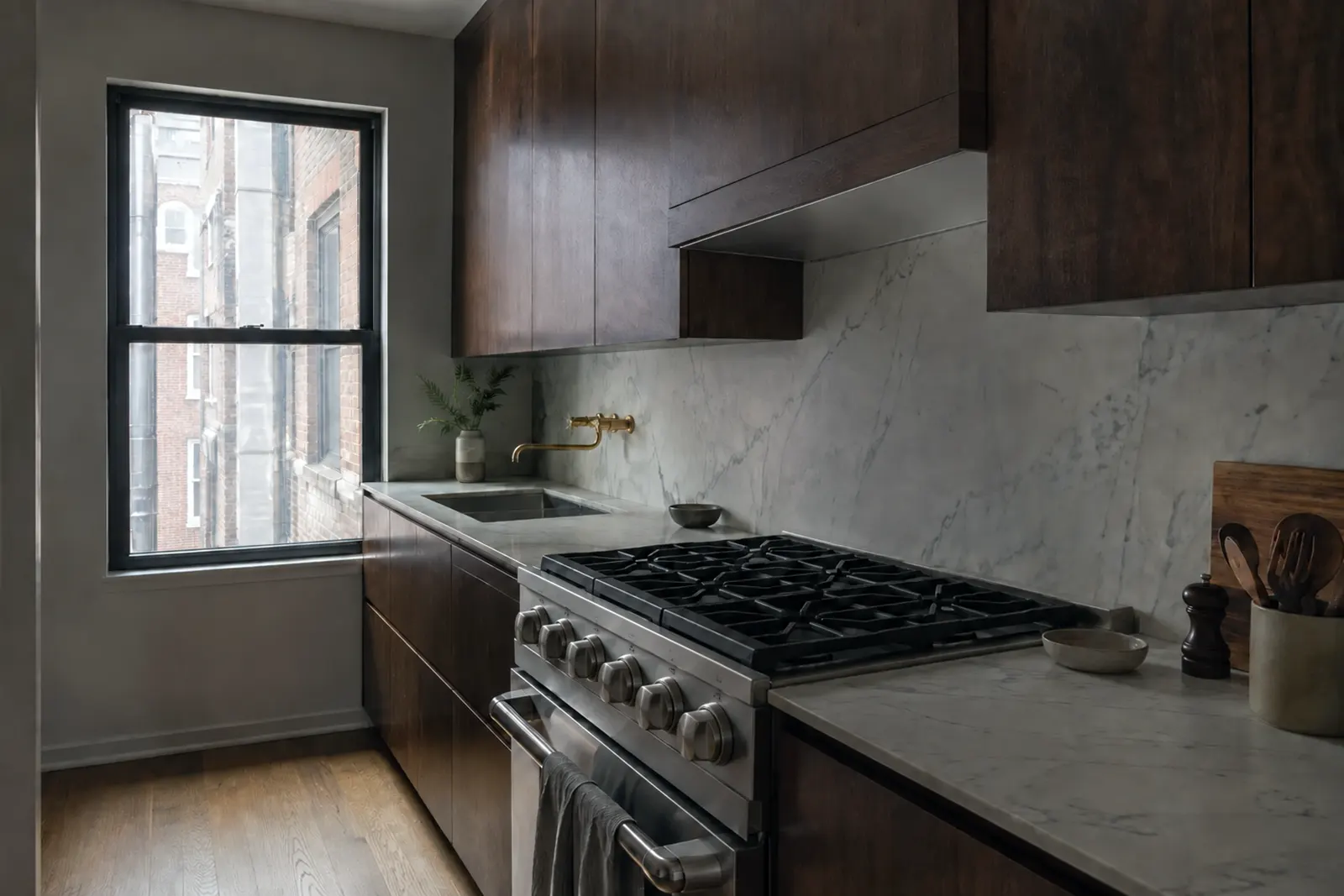 Dark walnut kitchen with honed white marble counter and backsplash, a stainless gas range under a walnut hood enclosure, a wall-mounted brass faucet over an undermount sink, and a window looking onto neighboring brick buildings