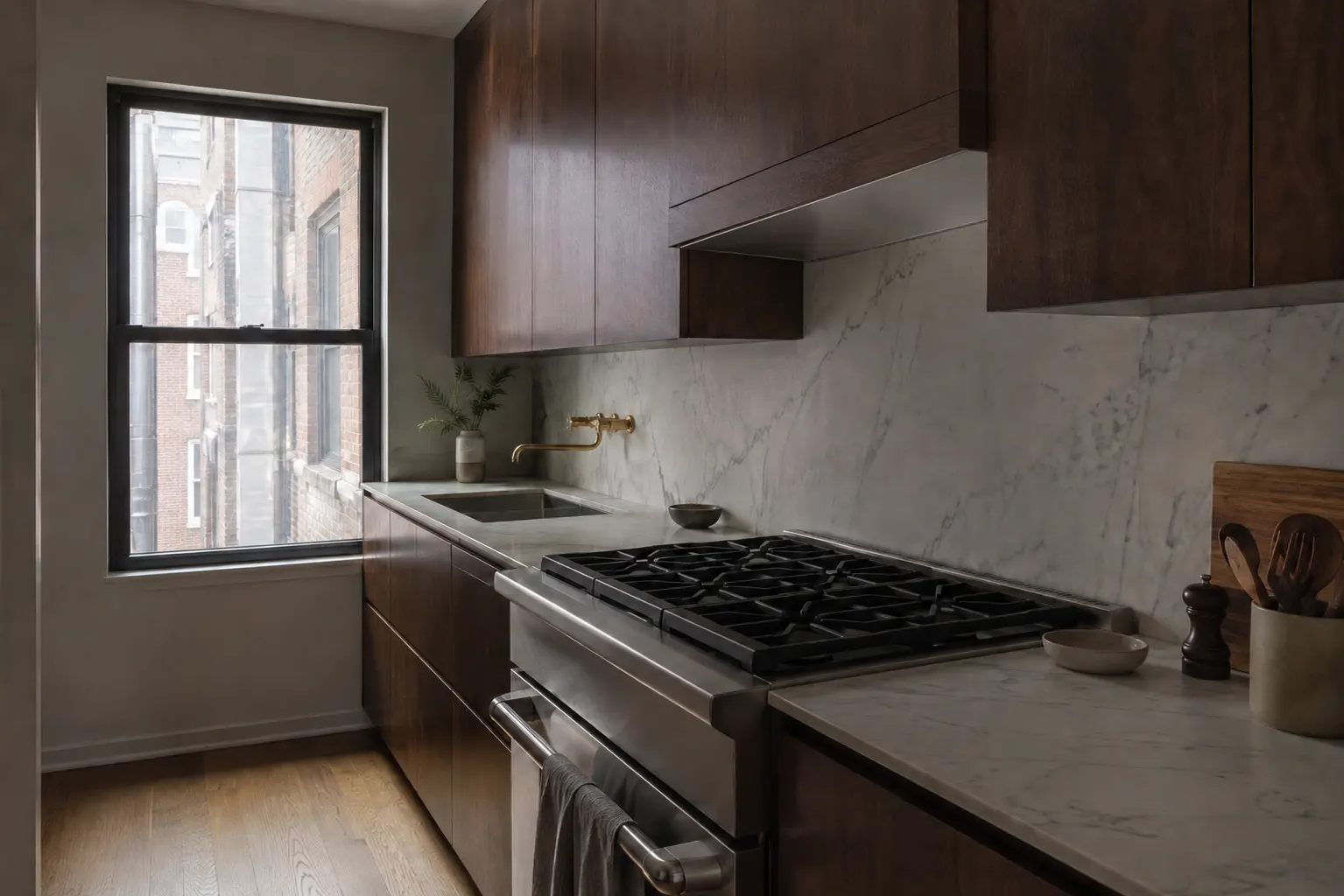 Dark walnut kitchen with honed white marble counter and backsplash, a stainless gas range under a walnut hood enclosure, a wall-mounted brass faucet over an undermount sink, and a window looking onto neighboring brick buildings